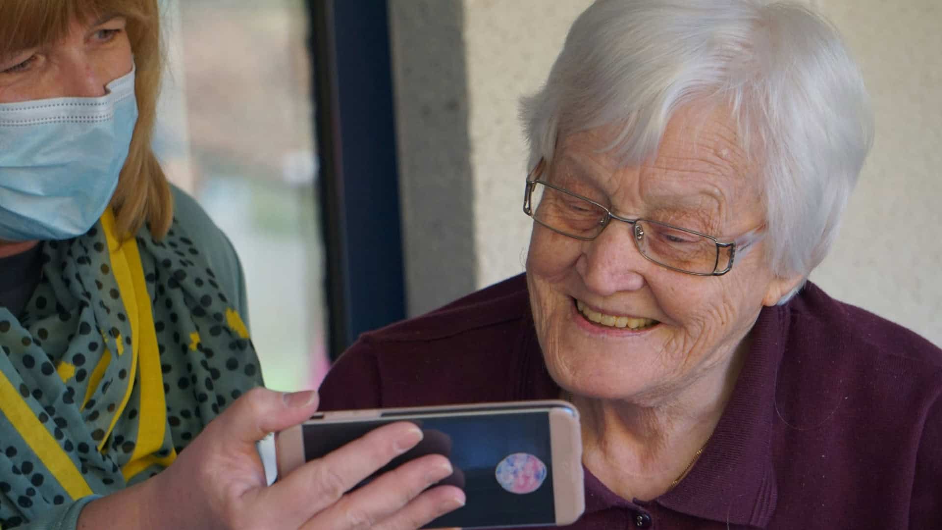 Pessoa idosa usando smartphone com apoio de outra pessoa, representando autonomia digital e inclusão.